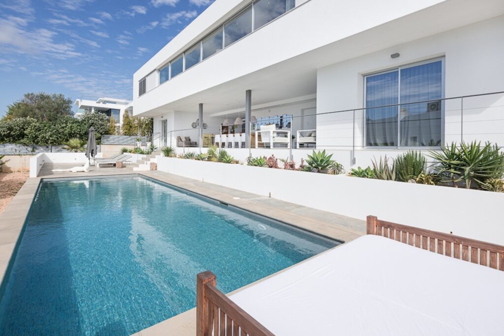 Villa in Vista Talamanca 6 Ibiza – Large Villa Rental Ibiza Main Description Large villa rental Ibiza, Villa in Vista Talamanca 6 Ibiza is a modern contemporary property located in the desirable village of Jesús, just minutes from Ibiza Town and Talamanca Beach. Designed with clean architectural lines and elegant finishes, this stylish villa offers privacy, comfort, and a prime location close to the beach. Prime Location Near Talamanca Beach & Ibiza Town Moreover, the villa is centrally positioned in Jesús, one of the most sought-after residential areas in Ibiza. As a result, guests can easily reach supermarkets, restaurants, and local services within minutes. At the same time, Talamanca Beach, Marina Botafoch, and Ibiza Town are only a short drive away, offering vibrant nightlife, exclusive restaurants, and luxury boutiques. Outdoor Living & Private Pool Area In addition, the villa features several covered and sun-drenched terraces distributed across different levels. The private swimming pool is located one level below the main living area and is surrounded by comfortable lounge beds. Furthermore, there is a covered outdoor dining terrace with a BBQ and outdoor bar, creating the perfect environment for relaxed afternoons and evening gatherings. The rooftop terrace offers additional space to enjoy open countryside views and the green landscape of Ibiza. Consequently, guests can choose between sun, shade, and panoramic views throughout the day. Contemporary Interiors & Functional Layout Inside, the villa combines velvet textures, wood finishes, and refined design elements. The open kitchen with a central cooking island flows seamlessly into the living and dining areas, creating a bright and welcoming space. Thanks to large windows and sliding doors, the interior connects naturally with the terraces and outdoor areas. The villa accommodates 8 guests across 4 bedrooms and 4 bathrooms, all designed with comfort and privacy in mind. Two bedrooms are located on the upper level, including the master suite with en-suite bathroom. The other two bedrooms are positioned on the lower level, each with independent entrances and en-suite bathrooms, providing additional privacy for guests. Overall, Villa in Vista Talamanca 6 Ibiza represents a refined large villa rental in Ibiza, ideal for families or small groups seeking modern comfort close to the beach and Ibiza Town. Accommodation & Rooms Sleeps up to 8 guests 4 bedrooms 4 bathrooms Master bedroom with en-suite bathroom and large closets Second bedroom with private bathroom Two lower-level bedrooms with independent entrances and en-suite bathrooms Air conditioning throughout Amenities & Features Private swimming pool Multiple terraces Rooftop terrace Covered outdoor dining area Outdoor bar & BBQ Fully equipped modern kitchen Sound system Satellite TV High-speed Wi-Fi Alarm system Laundry room Countryside views Private parking Location & Surroundings – Jesús & Talamanca, Ibiza Located in Jesús village Minutes from Talamanca Beach Close to Marina Botafoch Short drive to Ibiza Town Nearby supermarkets, restaurants, and gym Residential and family-friendly area Additional Services Available on Request To enhance your stay in Ibiza, we offer: Concierge services Private chef Extra cleaning Table service Massage and wellness treatments Nanny services Luxury car rental Yacht rental Private transfers All services can be arranged before arrival or during your stay. Villa Rental Rates (Per Week) Season Dates Price / Week Peak Season July & August € — High Season June € — Mid Season May & September € — Low Season April & October € — Minimum stay: 7 nights (Saturday – Saturday) Rental type: Weekly rental Technical Data & Specifications Property type: Modern contemporary villa Location: Jesús / Talamanca, Ibiza Guests: 8 Bedrooms: 4 Bathrooms: 4 Swimming pool: Private Air conditioning: Yes Internet: High-speed Wi-Fi Security: Alarm system Parking: Private Booking & Rental Information Welcome pack at arrival Cleaning: 2 hours per weekday + final cleaning Damage deposit: € 3,000 Booking policy: 50% down payment within 5 working days 50% one month before arrival Check-in: from 16:00 Check-out: before 10:00 No parties allowed